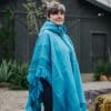 Model wearing turquoise fringed womens poncho in front of dark barn