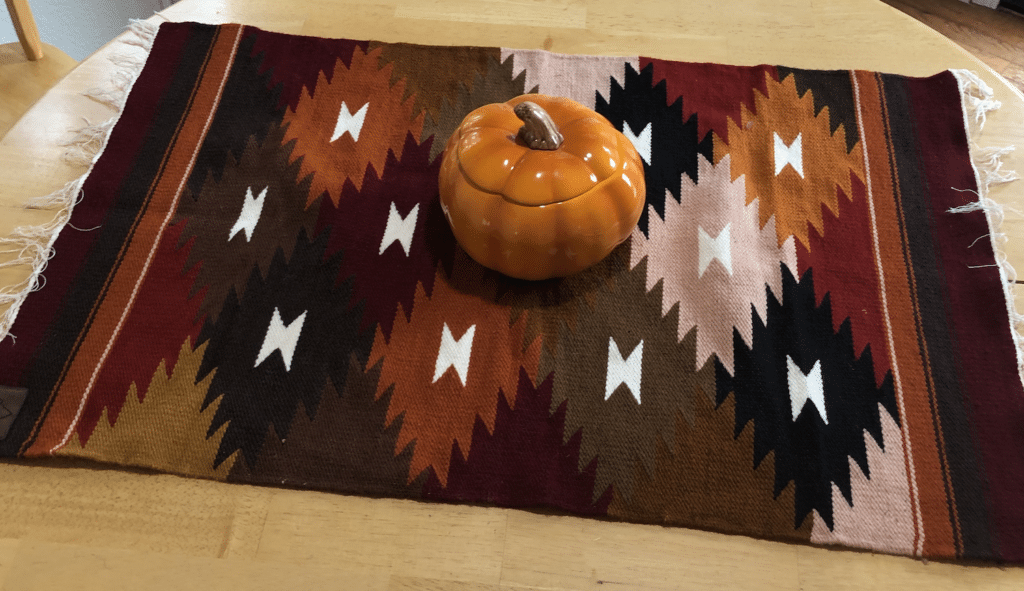 Handwoven wool tapestry with geometric shapes in earthtones on light oak table, with pumpkin-shaped soup tureen on top.