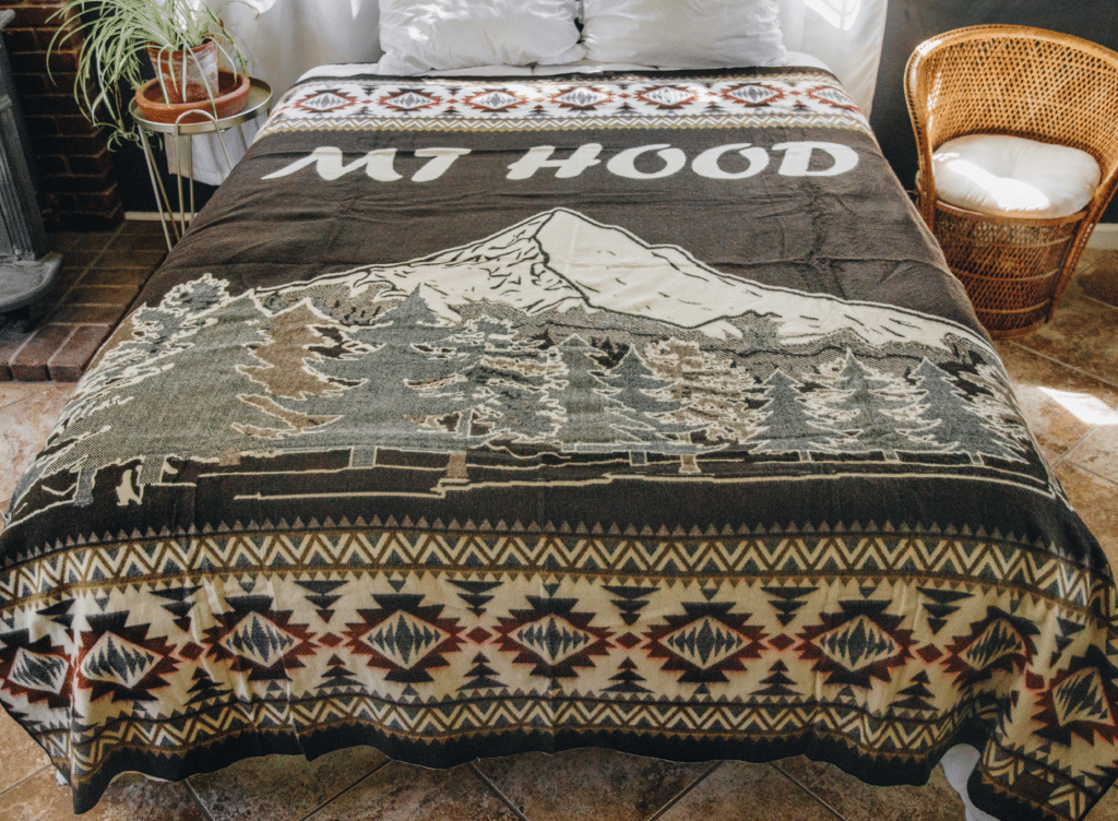 Brown blanket with mountain and text "Mt Hood" has geometric design on bottom and top edge. Displayed on a bed.