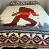 Blanket on bed has cream background, dark gray trees and large red-orange Bigfoot figure in center. Top and bottom edge have geometric design