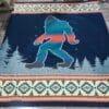 Blanket shows Bigfoot in turquoise with orange stripe on dark blue field, with small trees and geometric pattern across top and bottom