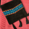 2 small square black suede crossbody bags with woven teal design, one with 3 tassels, one without, against red background