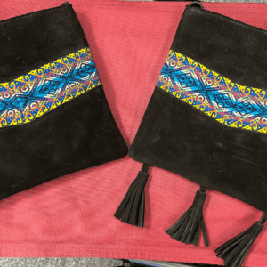 2 small square black suede crossbody bags with teal and yellow woven band design, one bag has 3 tassels, one has no tassels, laying on red background
