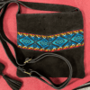 Small square black crossbody bag with adjustable strap and teal woven band, laying on red background