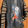 Military green hoodie with image of Bigfoot in watercolors on front, geometric patterned sleeves and kangaroo front pocket; hanging against a wooden fence