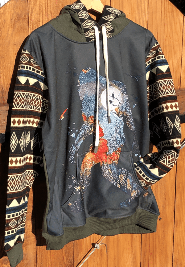 Military green hoodie with image of Bigfoot in watercolors on front, geometric patterned sleeves and kangaroo front pocket; hanging against a wooden fence